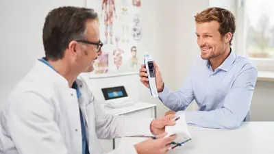 freundliches Arzt-Patientengespräch am Tisch, lächelnder Patient mit Vivatmo me in der rechten Hand, Vivatmo pro im Hintergrund.