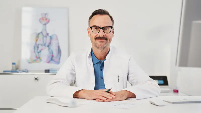 Confident-looking doctor sitting at desk in doctor's office environment with Vivatmo pro in background